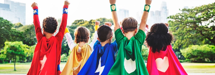 A group of children outdoors wearing colorful superhero capes, raising their arms in a gesture of empowerment and playfulness.